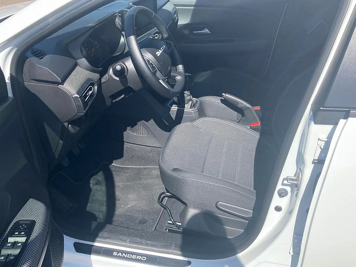 Vue intérieure côté conducteur de la Dacia Sandero blanche 2025, volant, siège tissu gris et levier de vitesse manuel visibles.