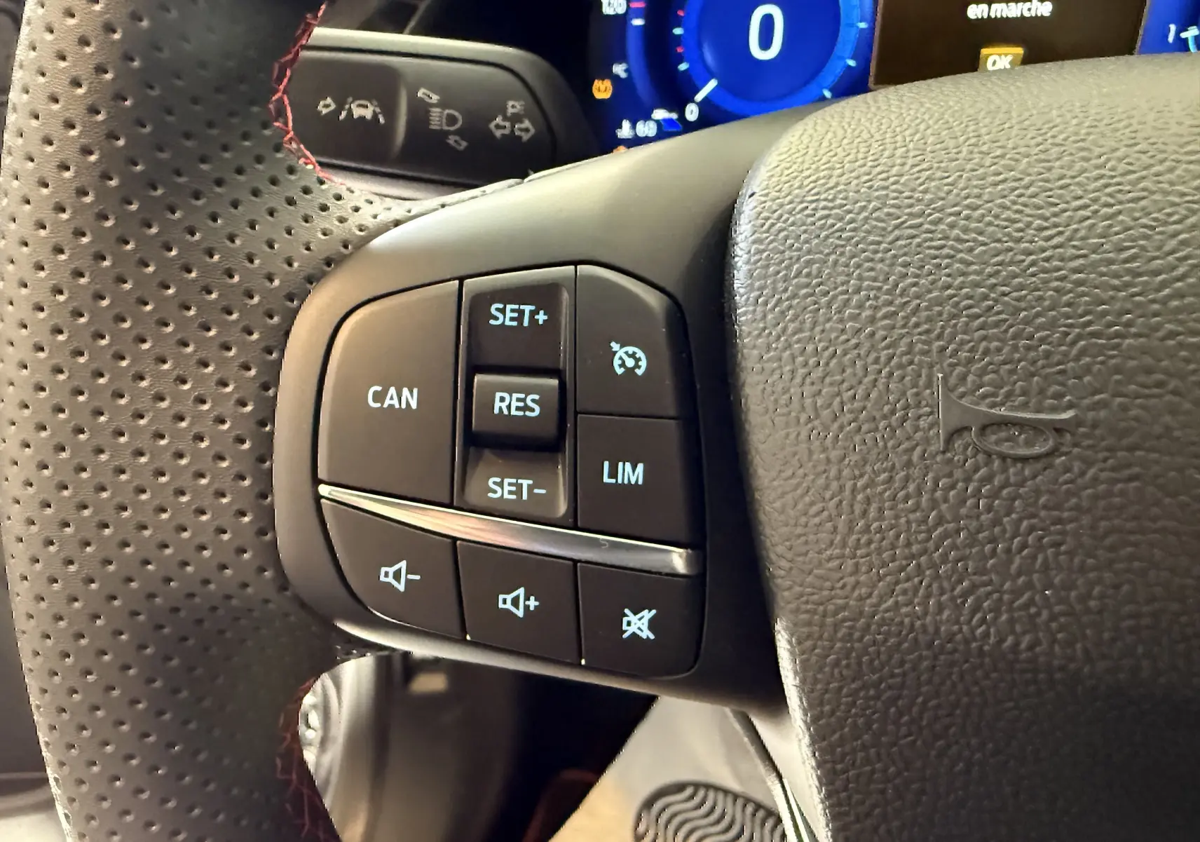 Gros plan sur le volant perforé avec commandes intégrées du côté gauche, tableau de bord numérique bleu en arrière-plan.