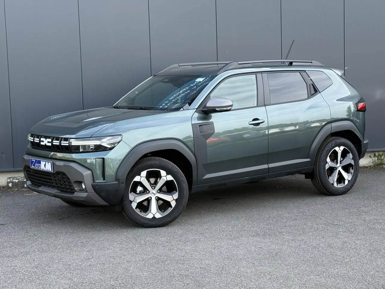 Vue 3/4 avant du Dacia Duster 2026 vert cèdre avec jantes alliage diamantées et barres de toit noires.