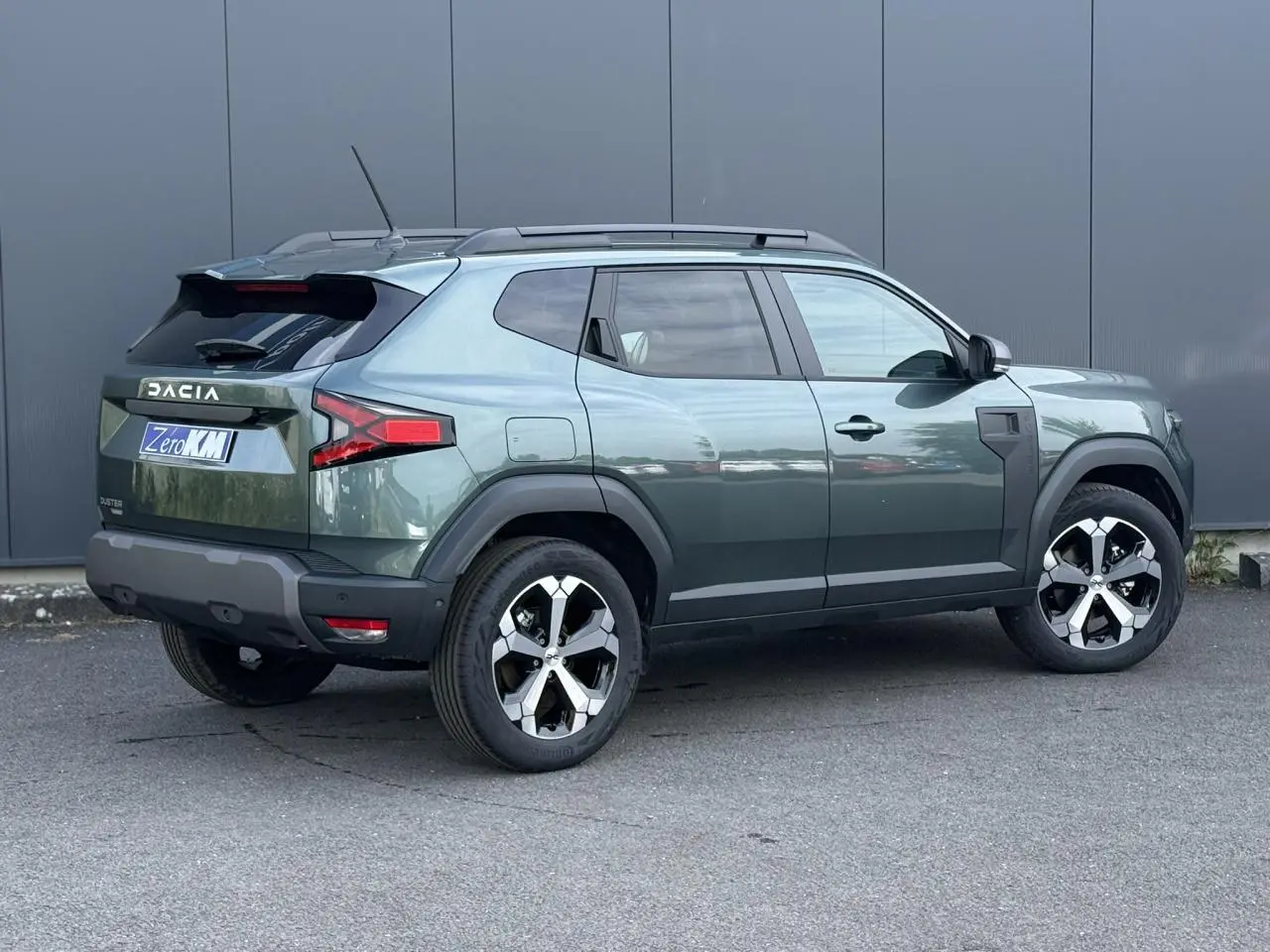 Vue 3/4 arrière droite du Dacia Duster 2026 vert cèdre avec jantes alliage diamantées et barres de toit noires.