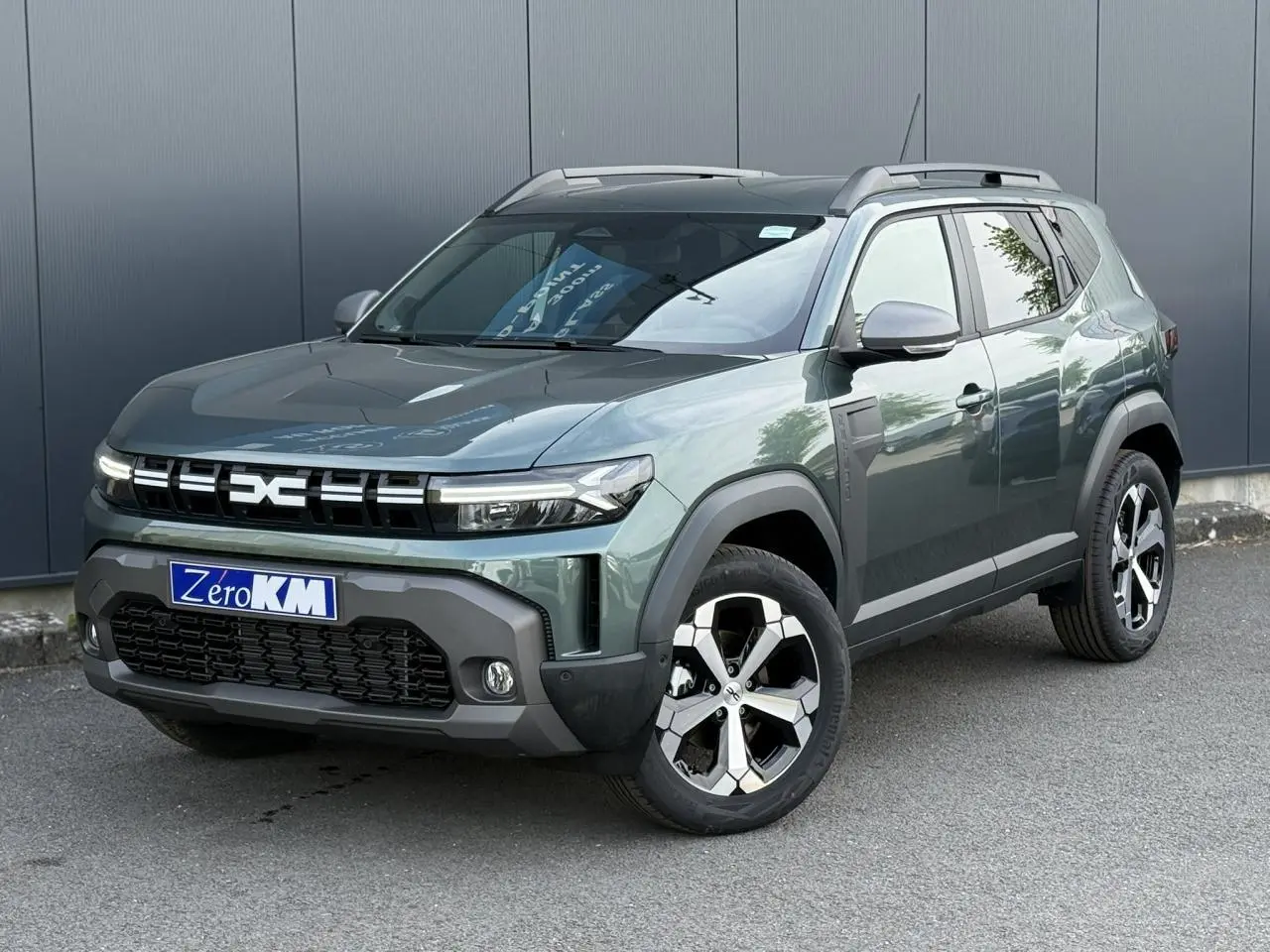 SUV Dacia Duster Vert Cèdre en 3/4 avant droit, avec jantes alliage et barres de toit noires.