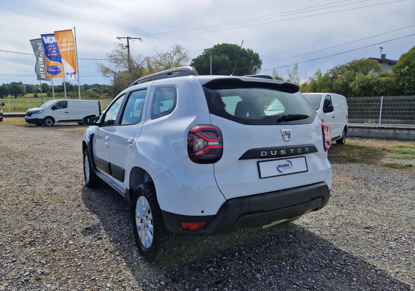 Vue 3/4 arrière droite d'un Dacia Duster blanc 2022 avec barres de toit et protections noires sur un terrain gravillonné.