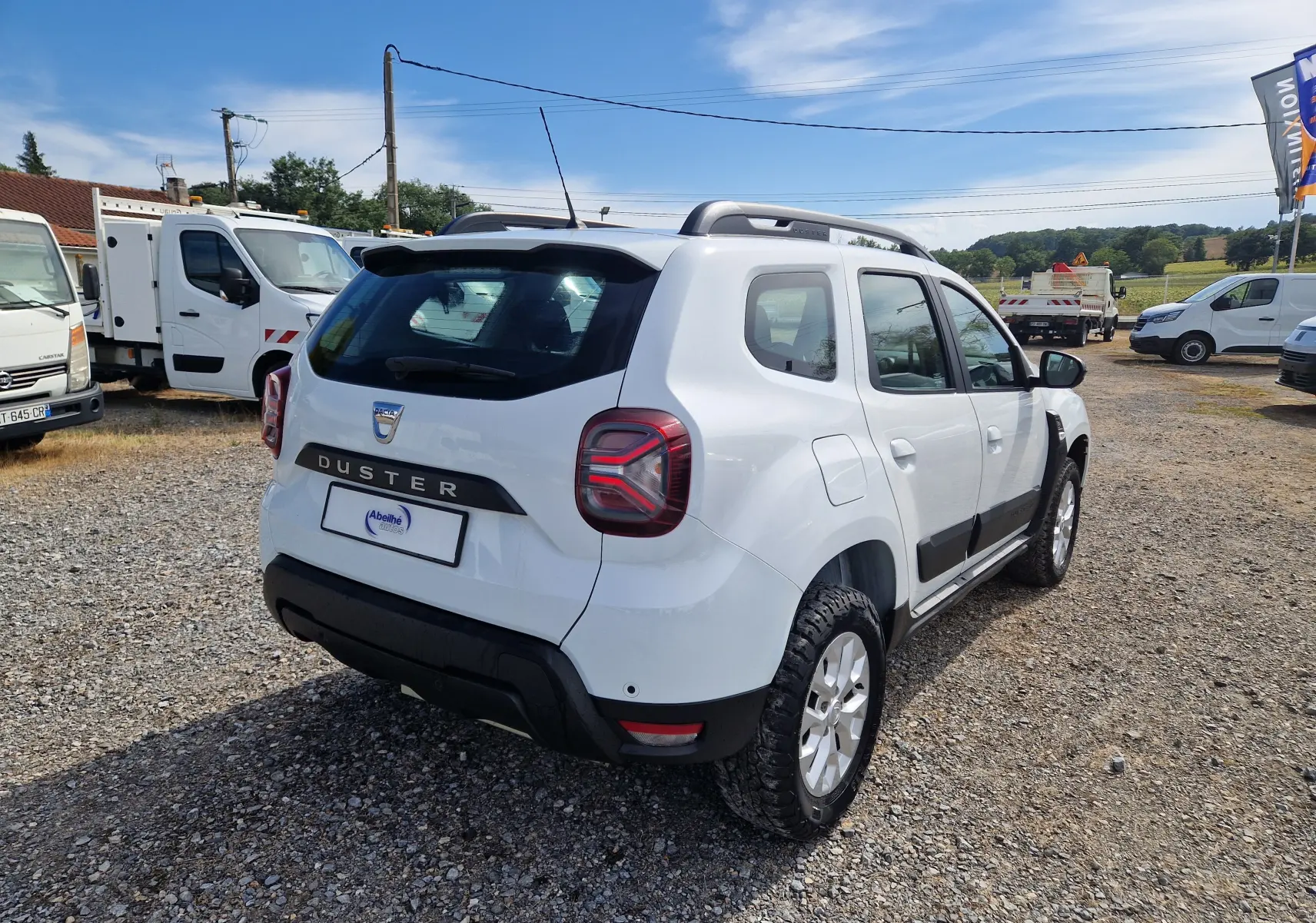 Vue 3/4 arrière droite d'un Dacia Duster blanc 4x4 2022 avec protections noires et jantes alliage sur un terrain gravillonné.