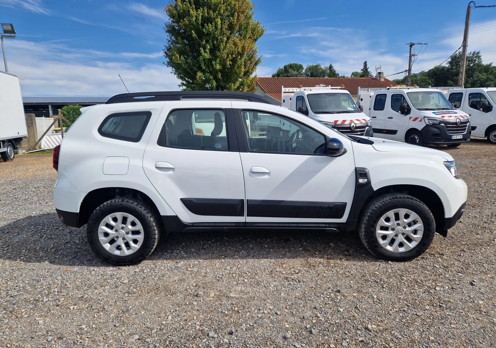 Profil côté gauche d'un Dacia Duster blanc 4x4 2022 avec protections noires et jantes alliage sur un sol gravillonné.