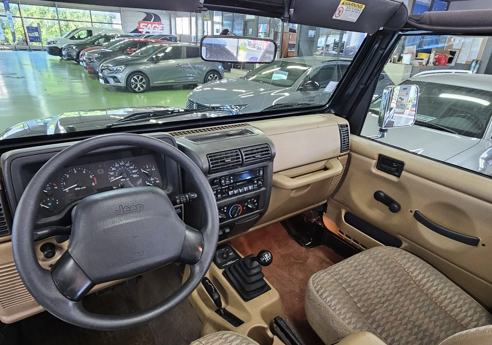 Intérieur beige d'une Jeep Wrangler noire de 2000, vue depuis le siège conducteur sur le tableau de bord et le levier de vitesses manuel.