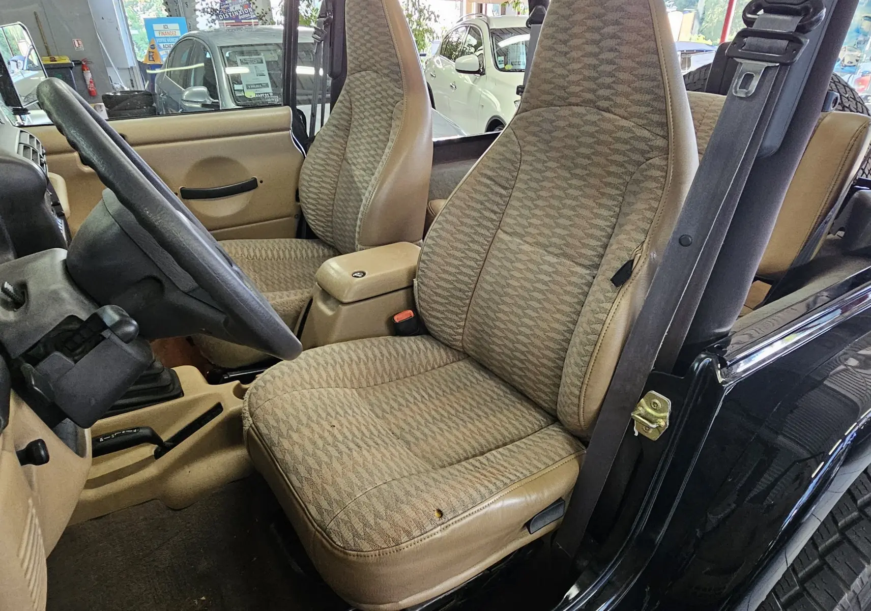 Intérieur beige du Jeep Wrangler 2000 vu côté conducteur, avec sièges en tissu à motifs et tableau de bord simple.