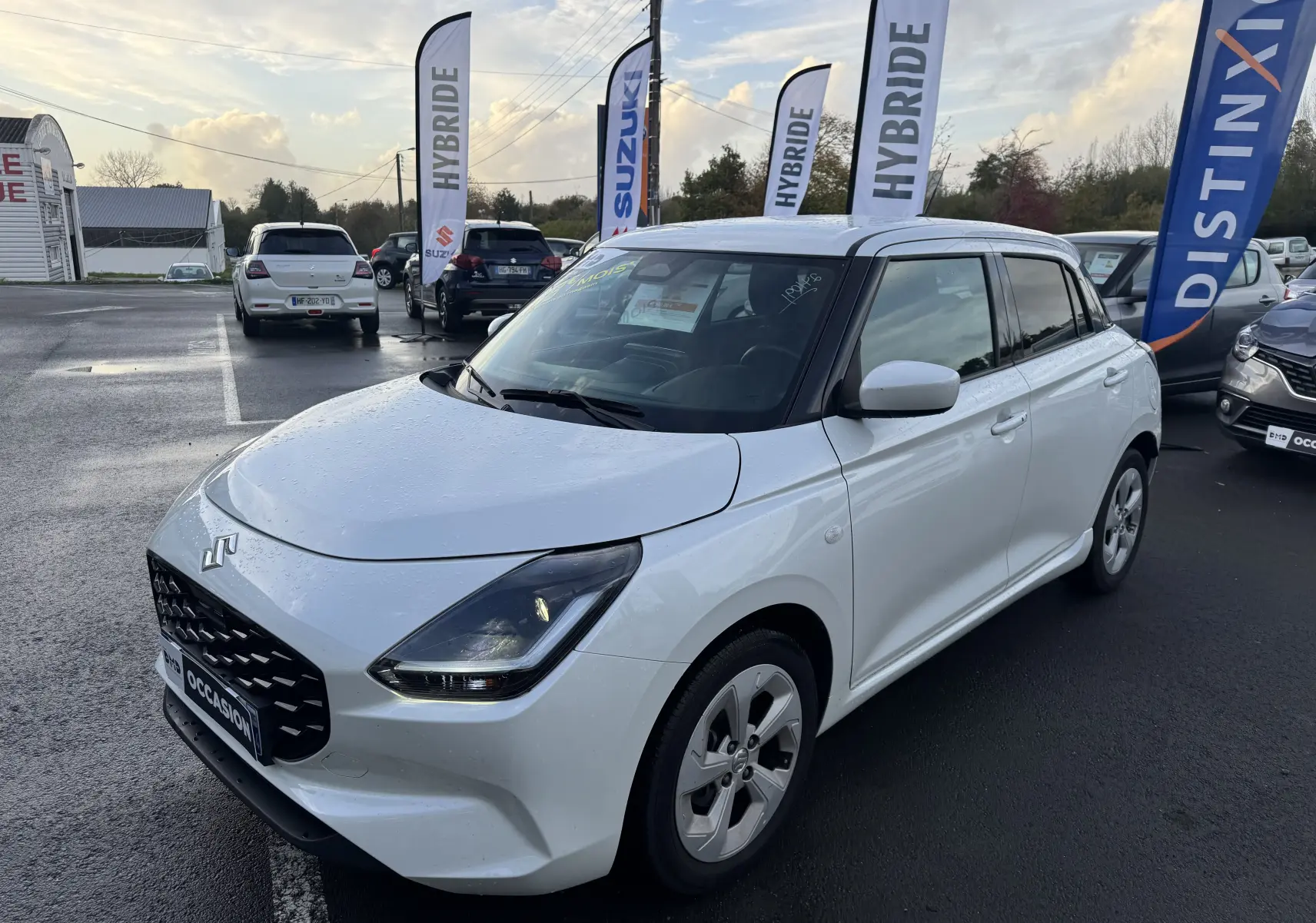 Suzuki Swift 1.2 Privilège blanc vue 3/4 avant droit avec calandre noire et feux LED allumés sur parking.