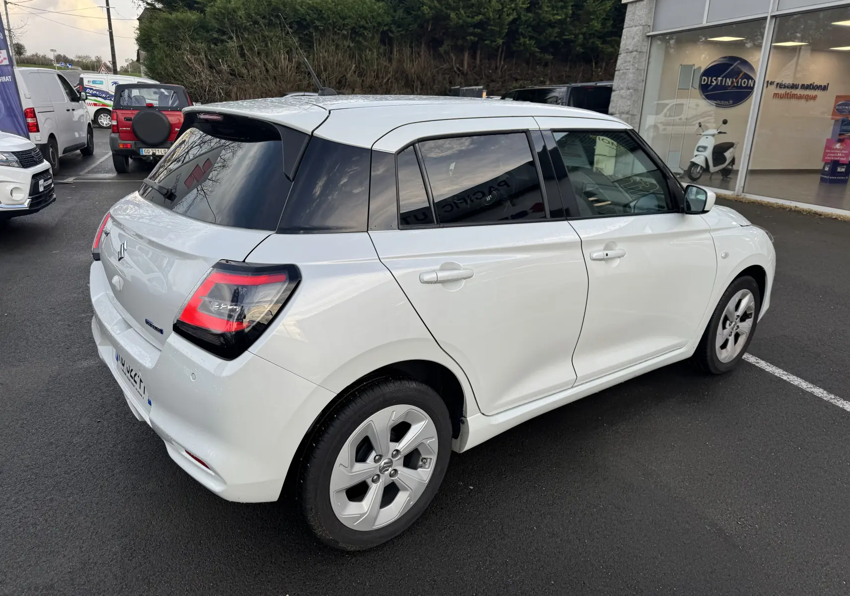 Suzuki Swift 1.2 Privilège blanc vue 3/4 arrière droit, avec vitres teintées et jantes spécifiques hybrides.