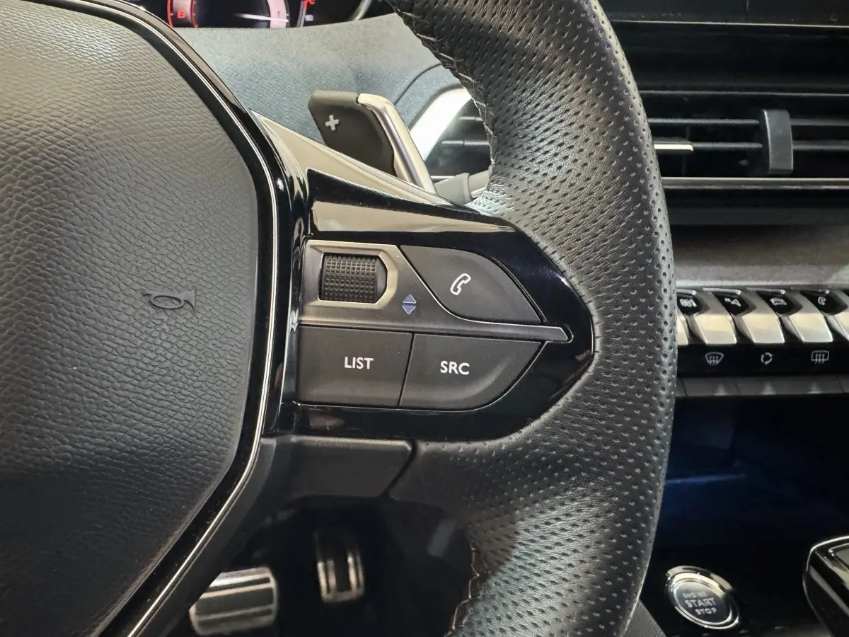 Gros plan sur le volant cuir perforé de la Peugeot 3008 GT, avec commandes audio et tableau de bord noir.