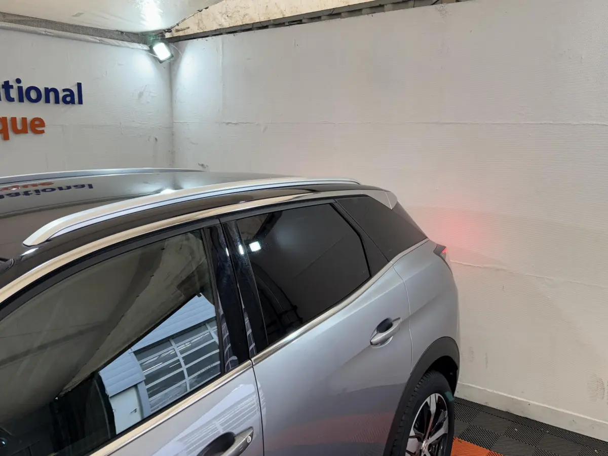 Vue 3/4 arrière droit du Peugeot 3008 GT gris clair avec vitres teintées et barres de toit chromées dans un garage.