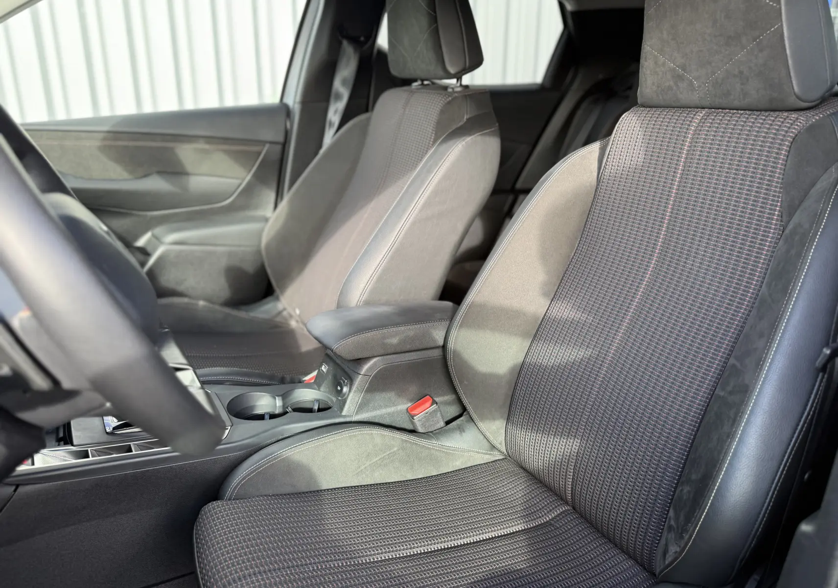 Vue intérieure côté avant gauche du DS3 hybride 2025, sièges en tissu gris avec surpiqûres rouges et accoudoir central.