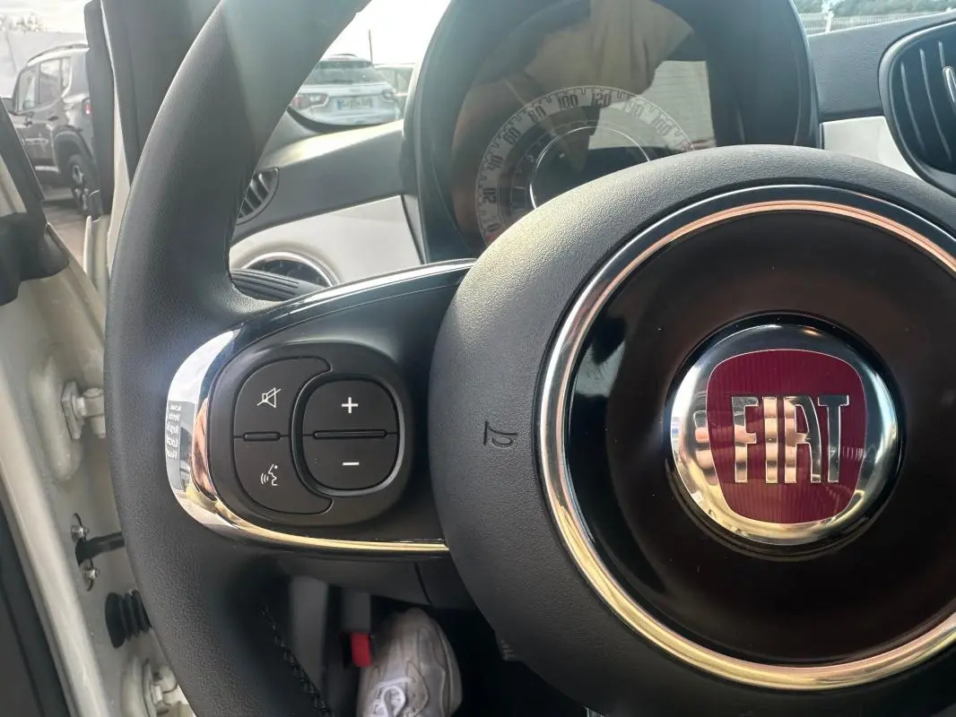 Gros plan sur le volant noir du Fiat 500 blanc 2023, avec logo FIAT rouge et commandes audio intégrées à gauche.