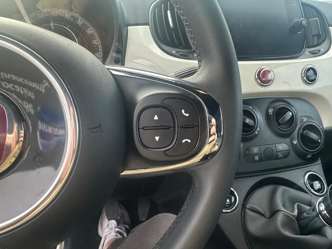 Vue intérieure centrée sur le volant de la Fiat 500 blanche 2023, avec commandes au volant et tableau de bord clair.