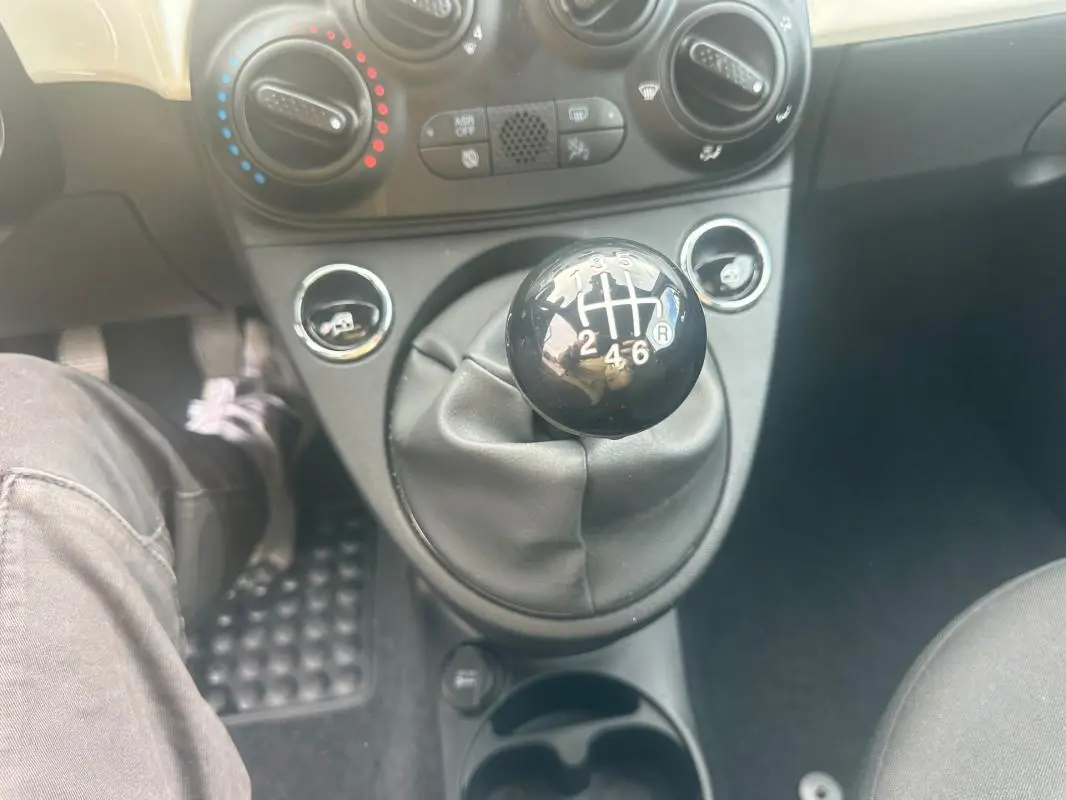 Gros plan sur le levier de vitesses manuel noir de la FIAT 500 blanche, avec commandes de climatisation en arrière-plan.
