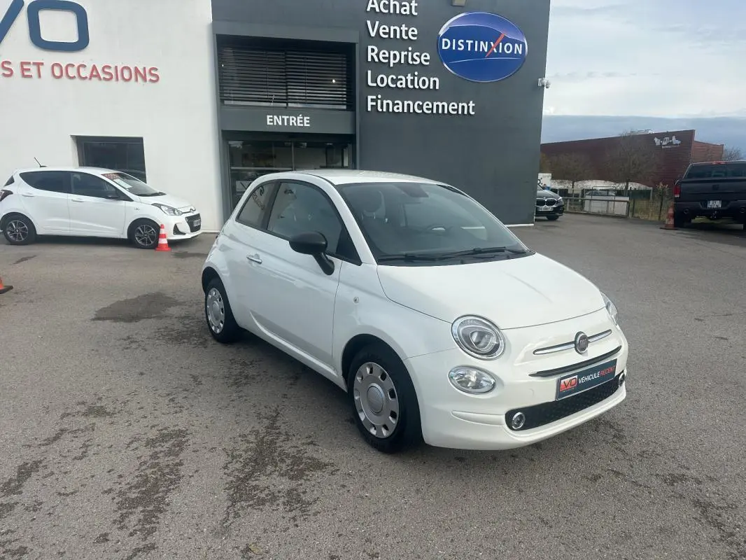 Vue 3/4 avant d'une FIAT 500 blanche de 2023, version hybride, avec rétroviseurs noirs, stationnée devant un bâtiment commercial.