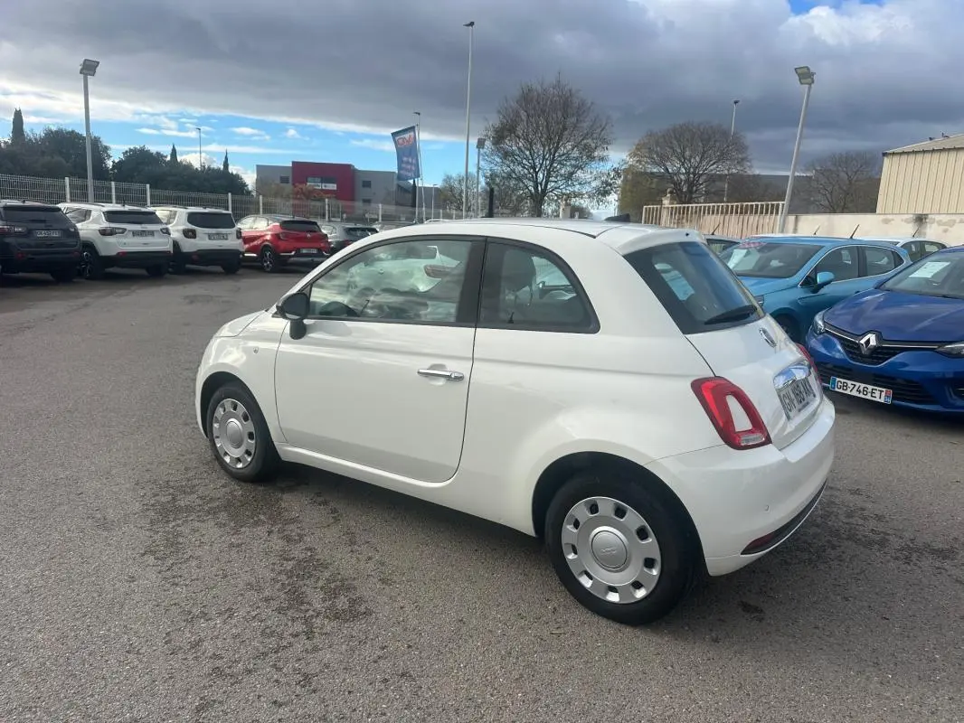 Vue 3/4 arrière droite d'une Fiat 500 blanche 2023, compacte et arrondie, stationnée en extérieur sur un parking.