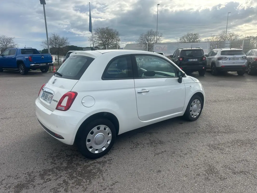Vue 3/4 arrière droite d'une FIAT 500 blanche de 2023, version hybride essence, garée en extérieur sur un parking.