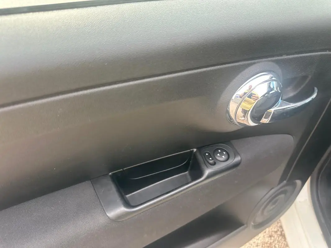 Vue rapprochée de la poignée intérieure et des commandes de vitre côté gauche d'une FIAT 500 blanche 2023.