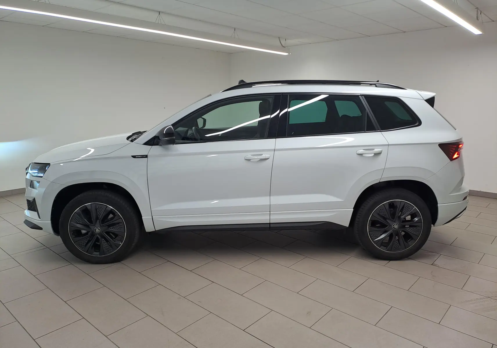 Vue de profil côté gauche d'un Skoda Karoq blanc lune 2025 avec jantes noires et toit ouvrant dans un showroom.