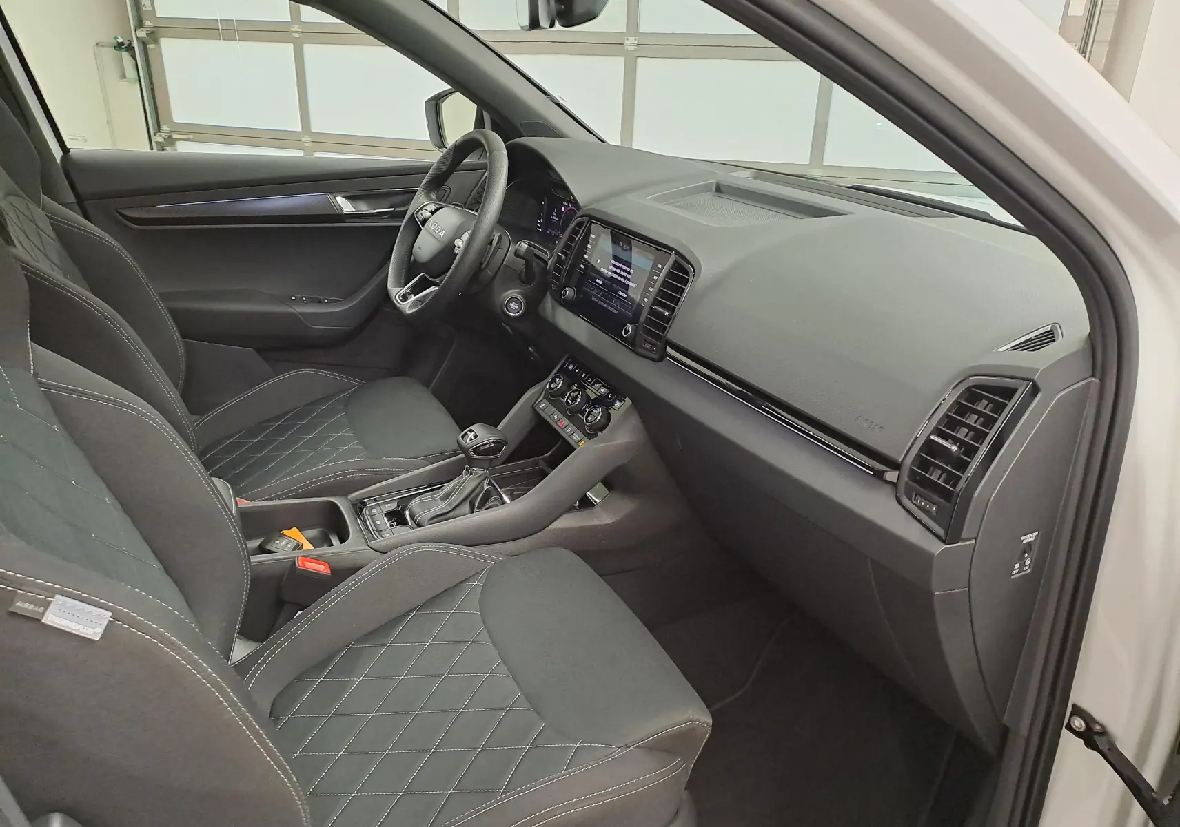 Intérieur du Skoda Karoq 2025 blanc lune, vue côté conducteur, sièges noirs avec surpiqûres et tableau de bord moderne.