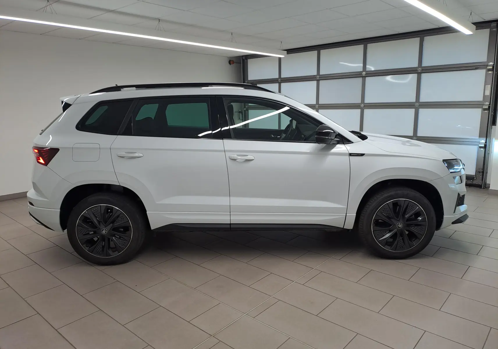 Vue de profil côté gauche d’un Skoda Karoq Blanc Lune 2025 avec jantes noires et toit noir en intérieur showroom.