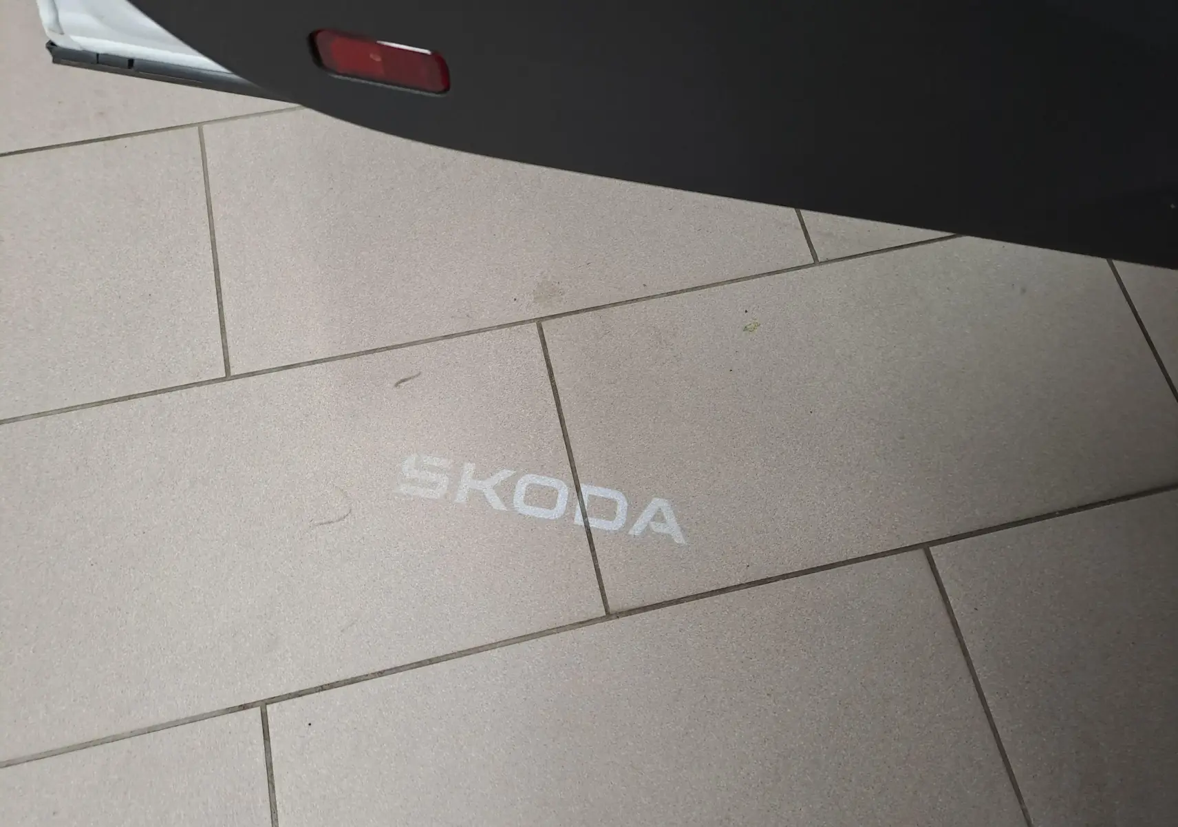 Projection lumineuse blanche du logo SKODA au sol, vue partielle de la porte ouverte d'un SKODA KAROQ Blanc Lune.