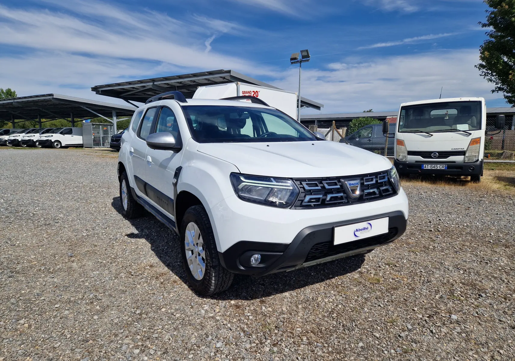 Dacia Duster 4x4 blanc en 3/4 avant droit sur parking extérieur, avec calandre noire et jantes alliage 16 pouces.