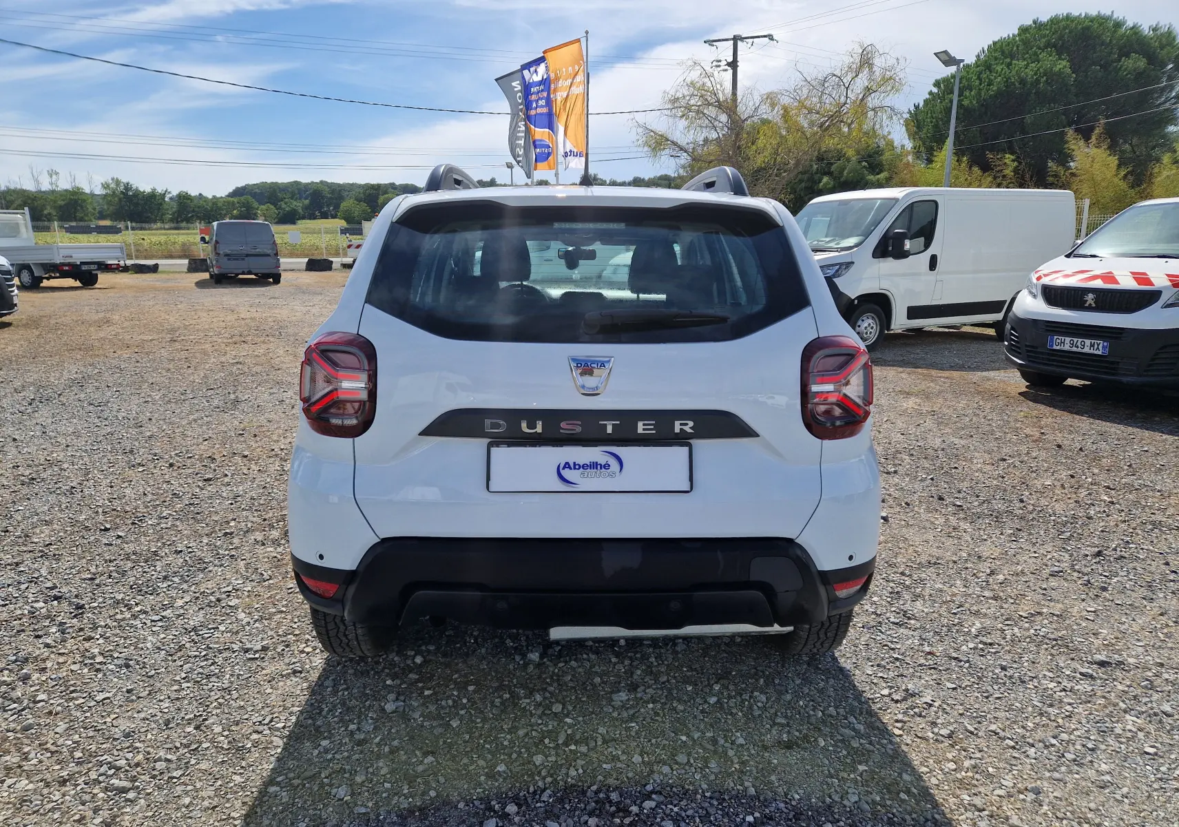 Vue arrière d'un Dacia Duster blanc 2023, version 4x4, sur un parking extérieur avec plusieurs véhicules autour.