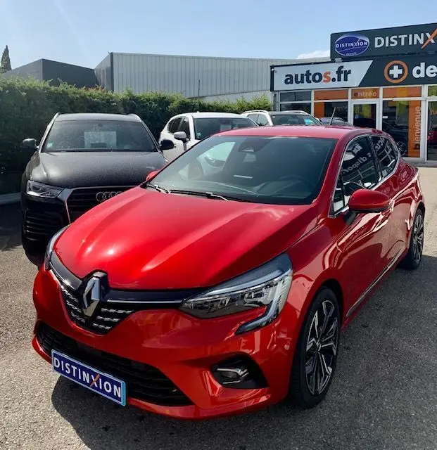 Renault Clio V rouge vue 3/4 avant droit, avec calandre distinctive et jantes alliage, stationnée devant un concessionnaire.