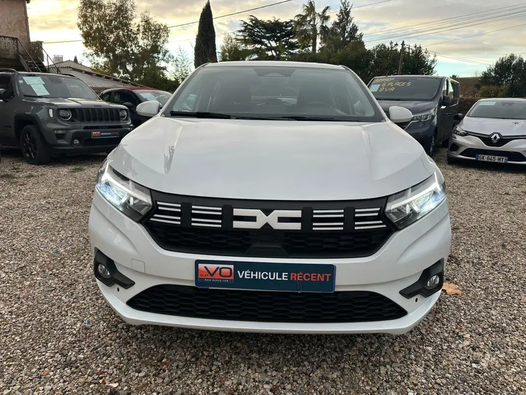 Vue de face d'une Dacia Sandero TCe 90 blanche avec calandre noire et phares allumés sur parking extérieur.