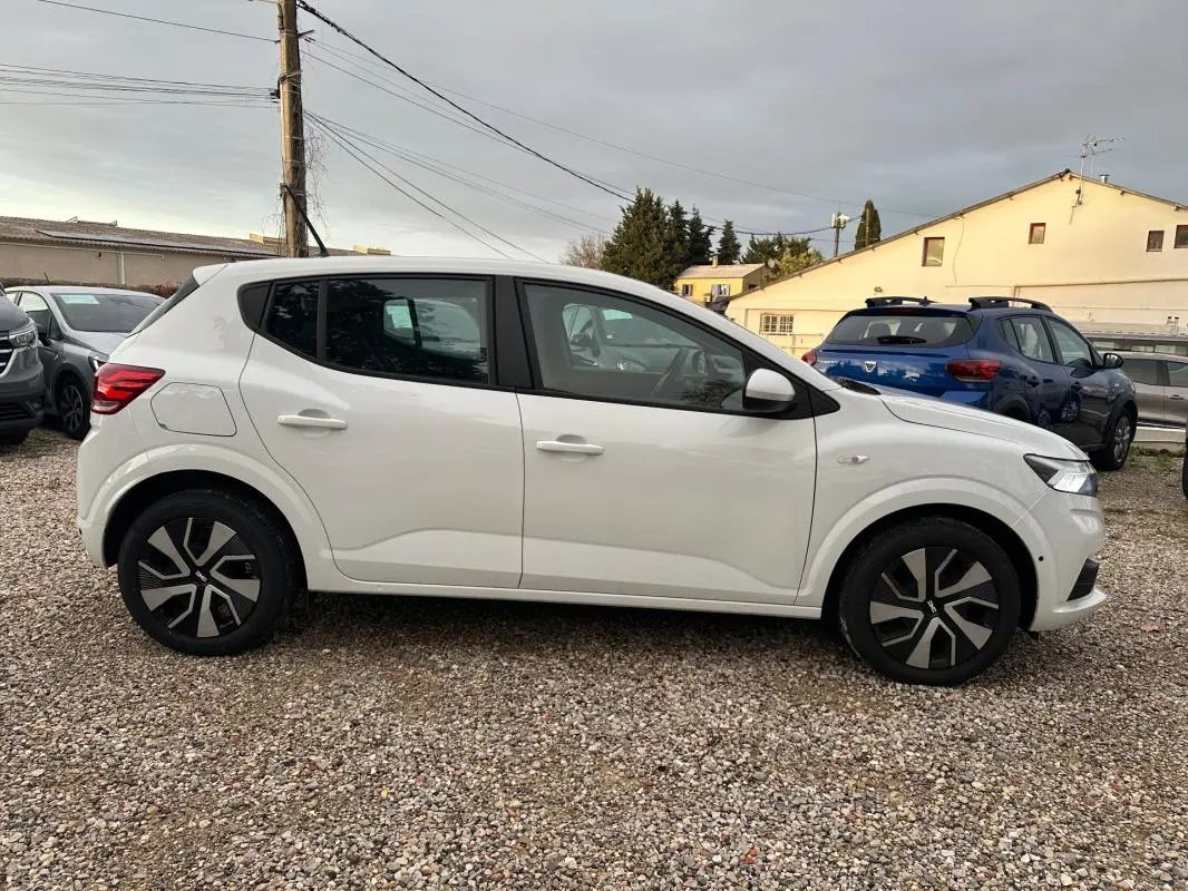 Vue de profil côté gauche d'une Dacia Sandero blanche 2024 garée sur un sol gravillonné, avec jantes noires et grises.