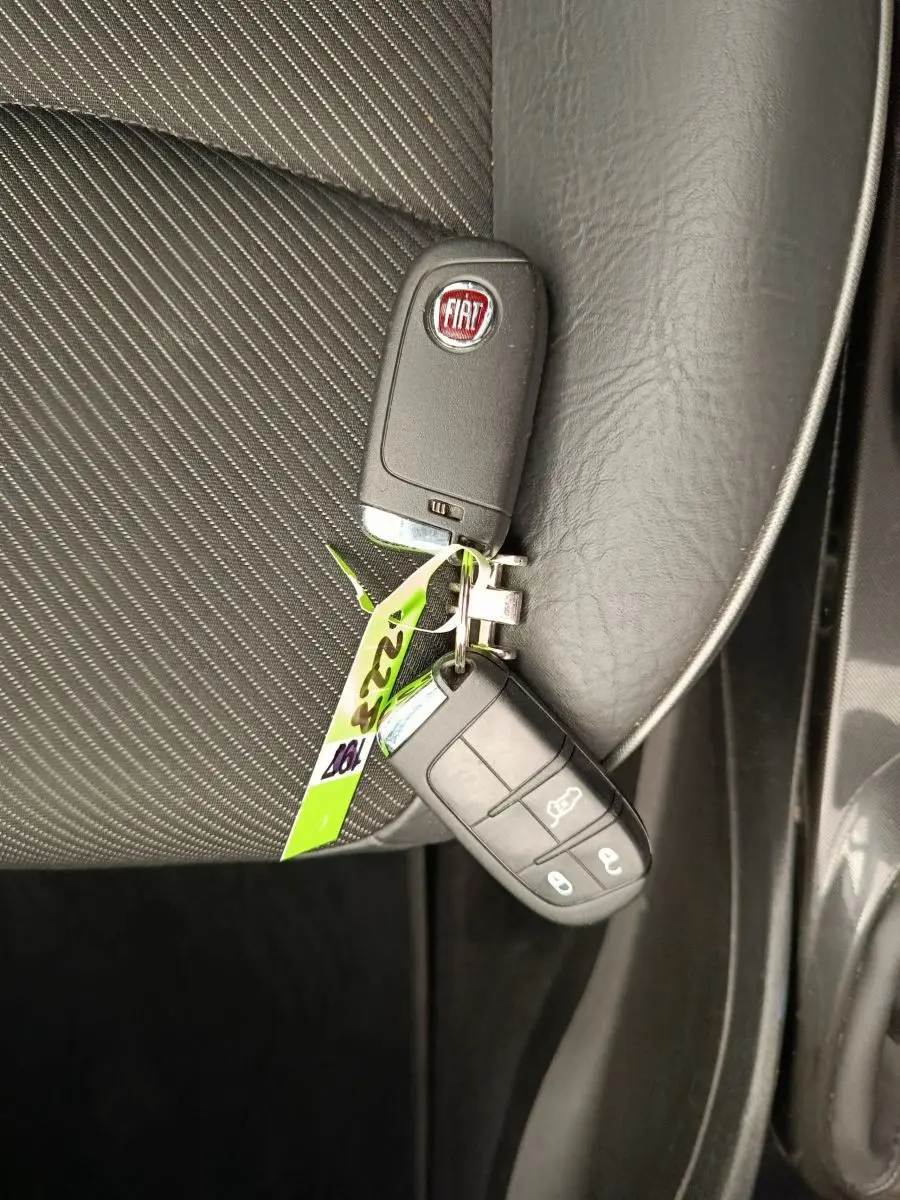 Clé FIAT posée sur siège tissu noir à coutures blanches, vue en plongée côté passager avant.