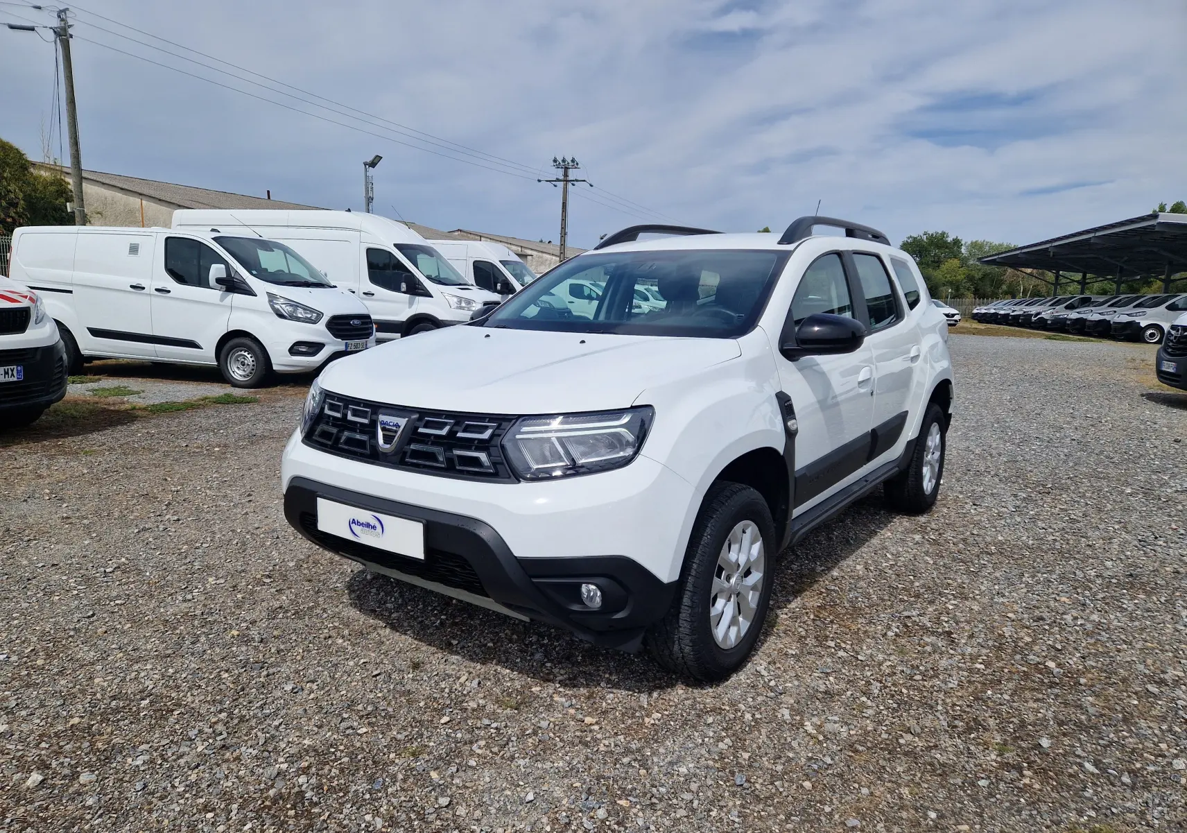 Dacia Duster blanc vue 3/4 avant droit sur parking gravillonné avec protections noires et barres de toit.