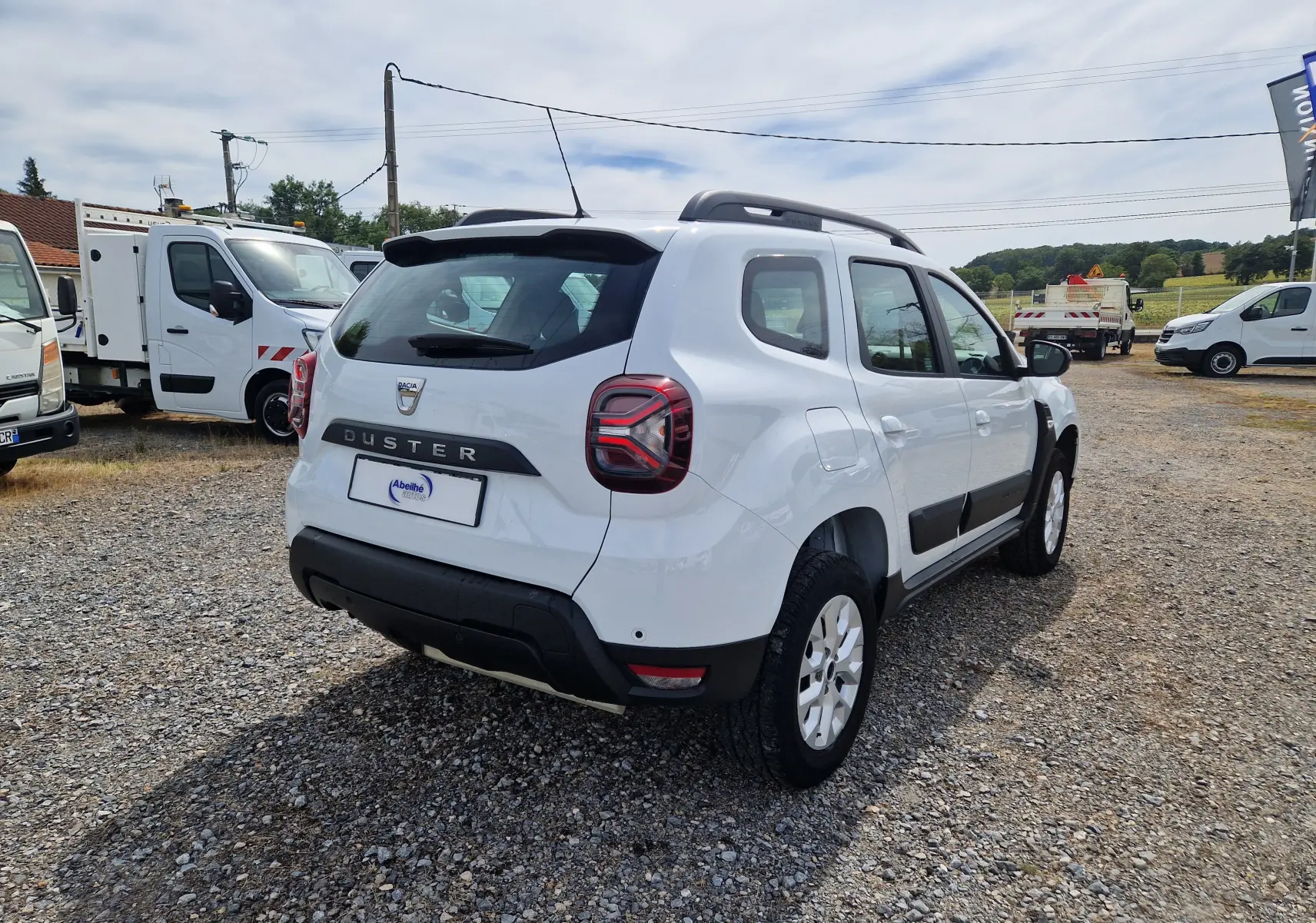Vue 3/4 arrière droite d'un Dacia Duster blanc 4x4 avec barres de toit et protections noires sur un sol gravillonné.