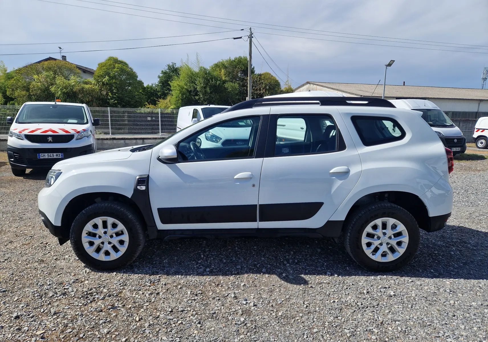 Vue de profil côté gauche d'un Dacia Duster 4x4 blanc 2023 avec protections noires et jantes 16 pouces.