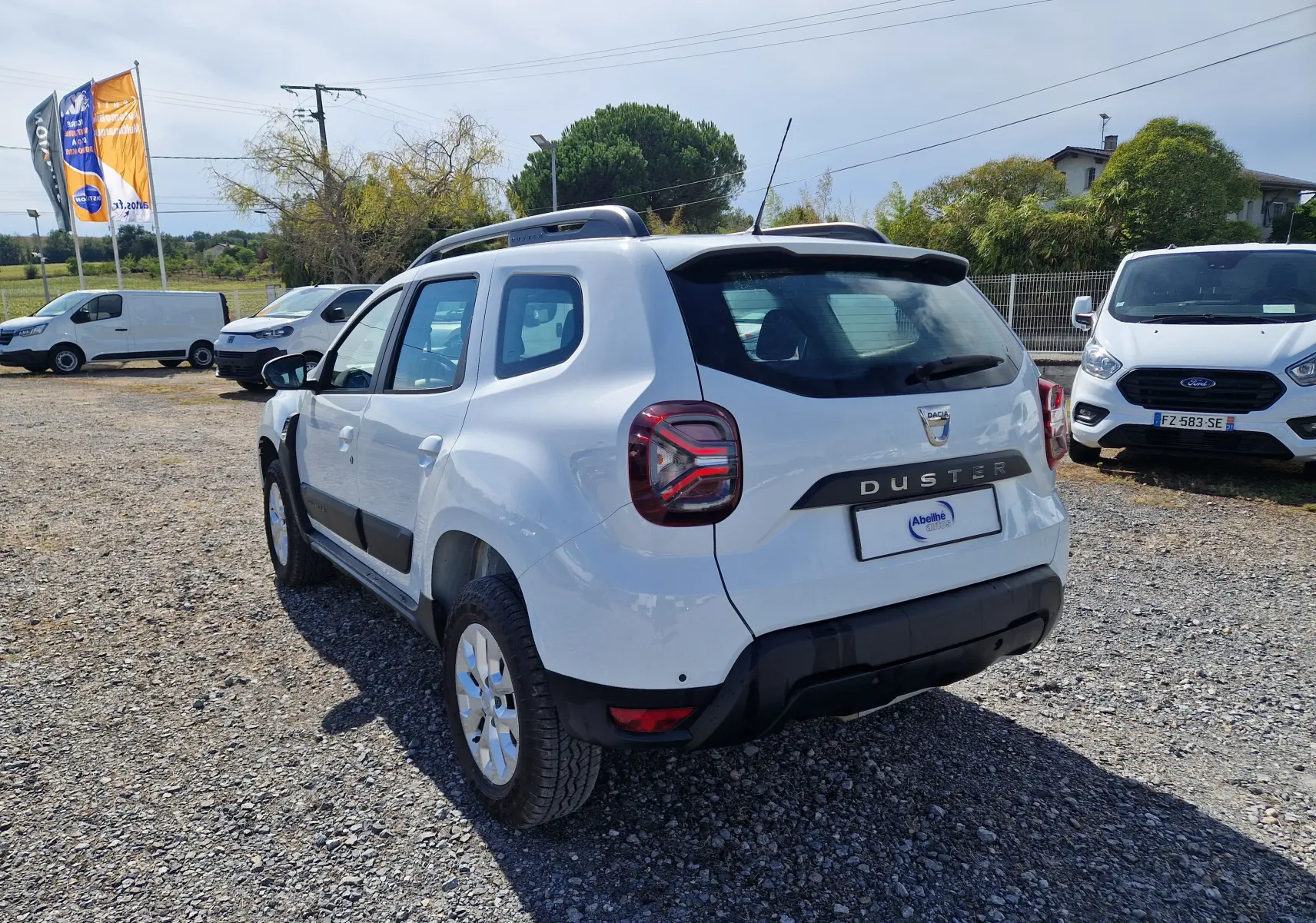 Vue 3/4 arrière droite d'un Dacia Duster blanc 2023 avec barres de toit et protections noires sur un terrain gravillonné.