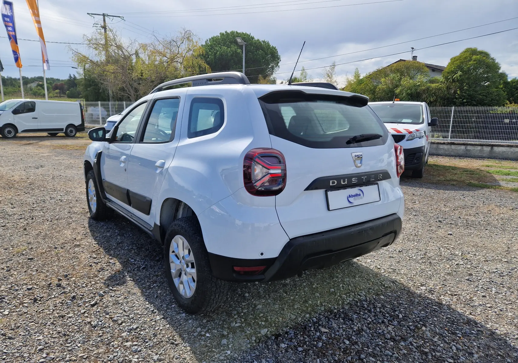 Vue 3/4 arrière droite du Dacia Duster blanc 2023 avec barres de toit et protections noires sur les portes.