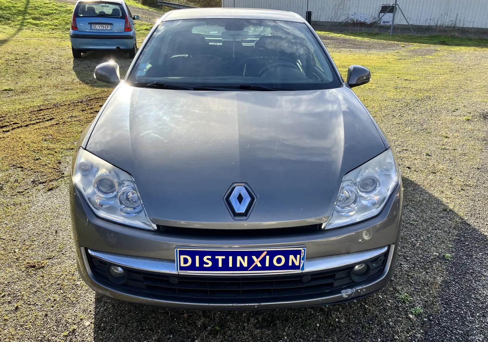 Vue frontale d'une Renault Laguna gris vert 2008 avec phares clairs et plaque d'immatriculation Distinxion visible.