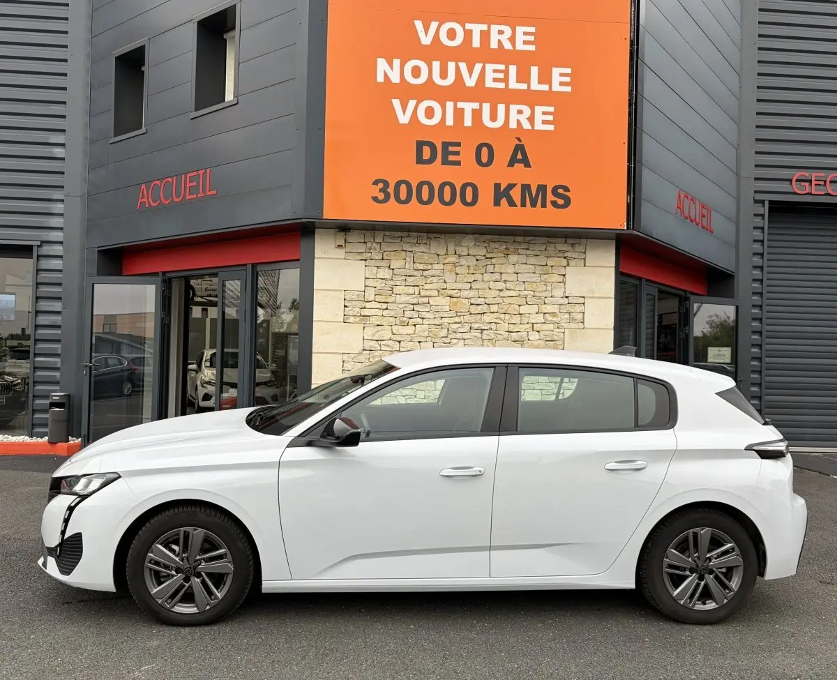 Profil droit d'une Peugeot 308 blanche 2024 garée devant un bâtiment avec enseigne orange "Votre nouvelle voiture de 0 à 30000 kms".