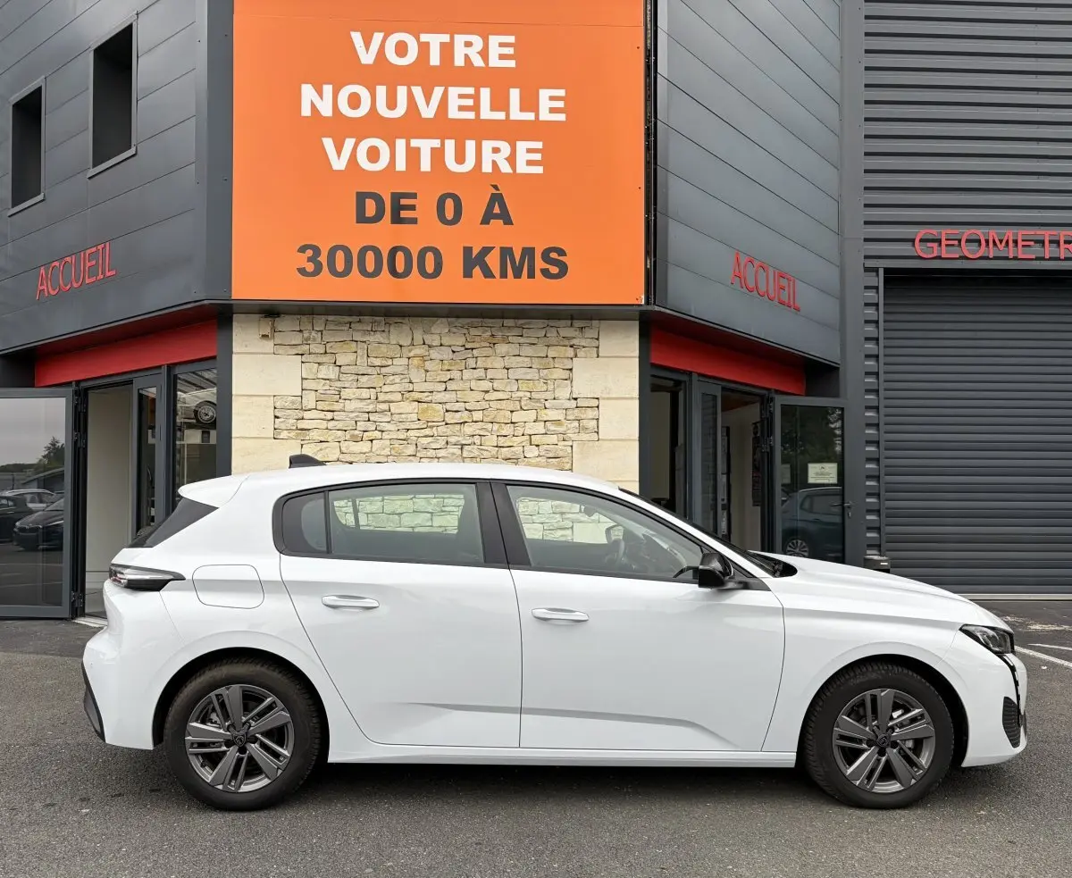 Profil latéral droit d'une Peugeot 308 blanche 2024 garée devant un bâtiment avec une grande enseigne orange.