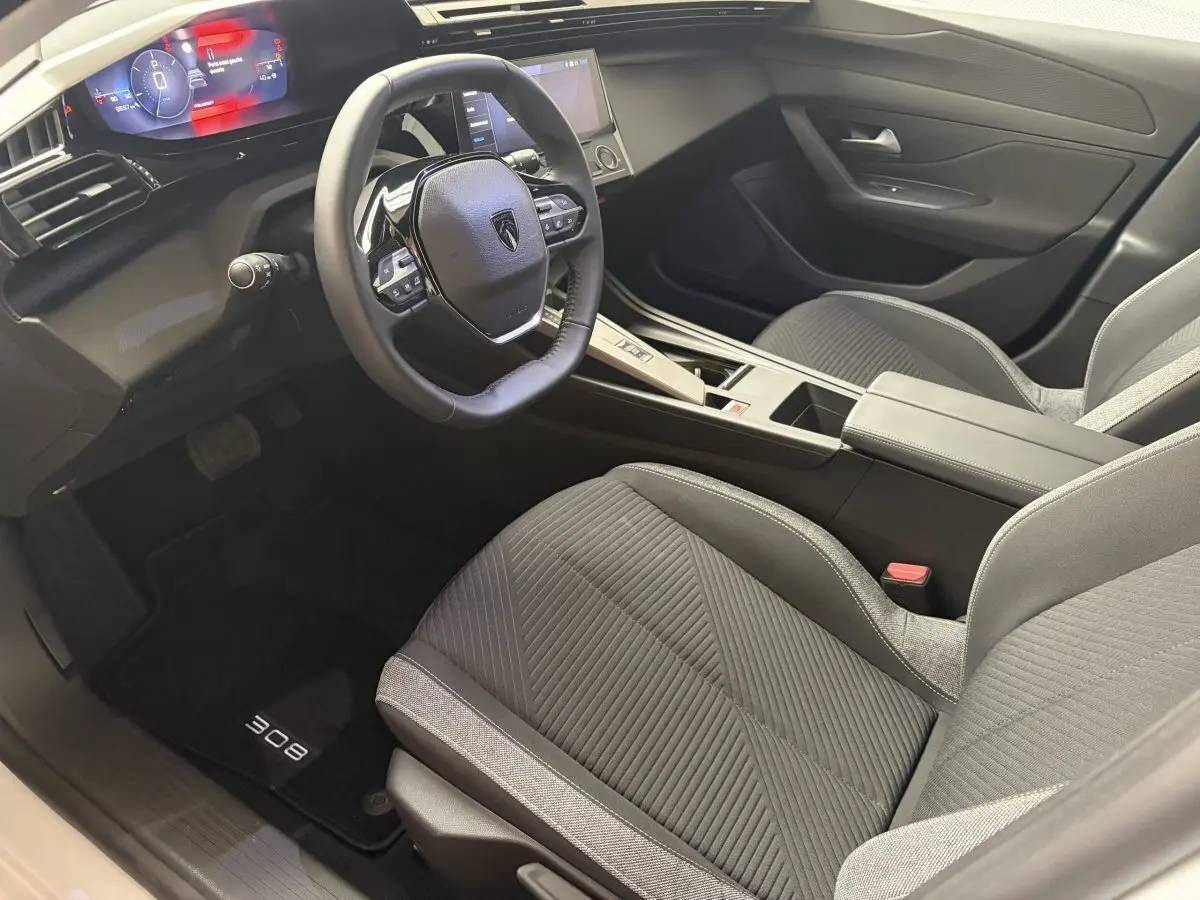 Intérieur de la Peugeot 308 2024 vu côté conducteur, sièges gris, volant cuir multifonctions et tableau de bord digital.