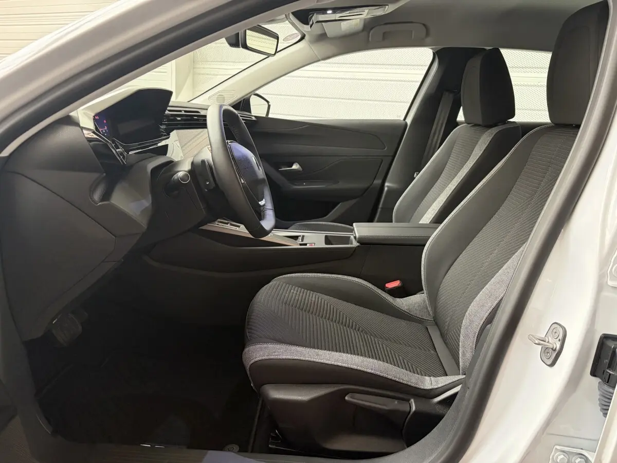 Intérieur avant de la Peugeot 308 blanche 2024, vue côté conducteur, sièges tissu noir et gris, tableau de bord moderne.