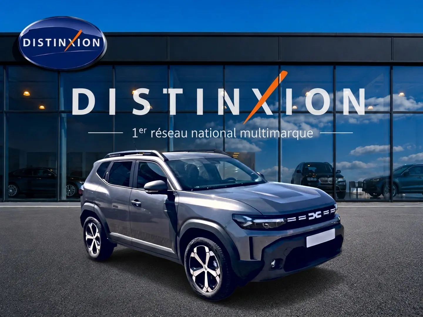 Dacia Duster gris schiste en 3/4 avant droit, avec barres de toit et jantes alu visibles devant un showroom Distinxion.