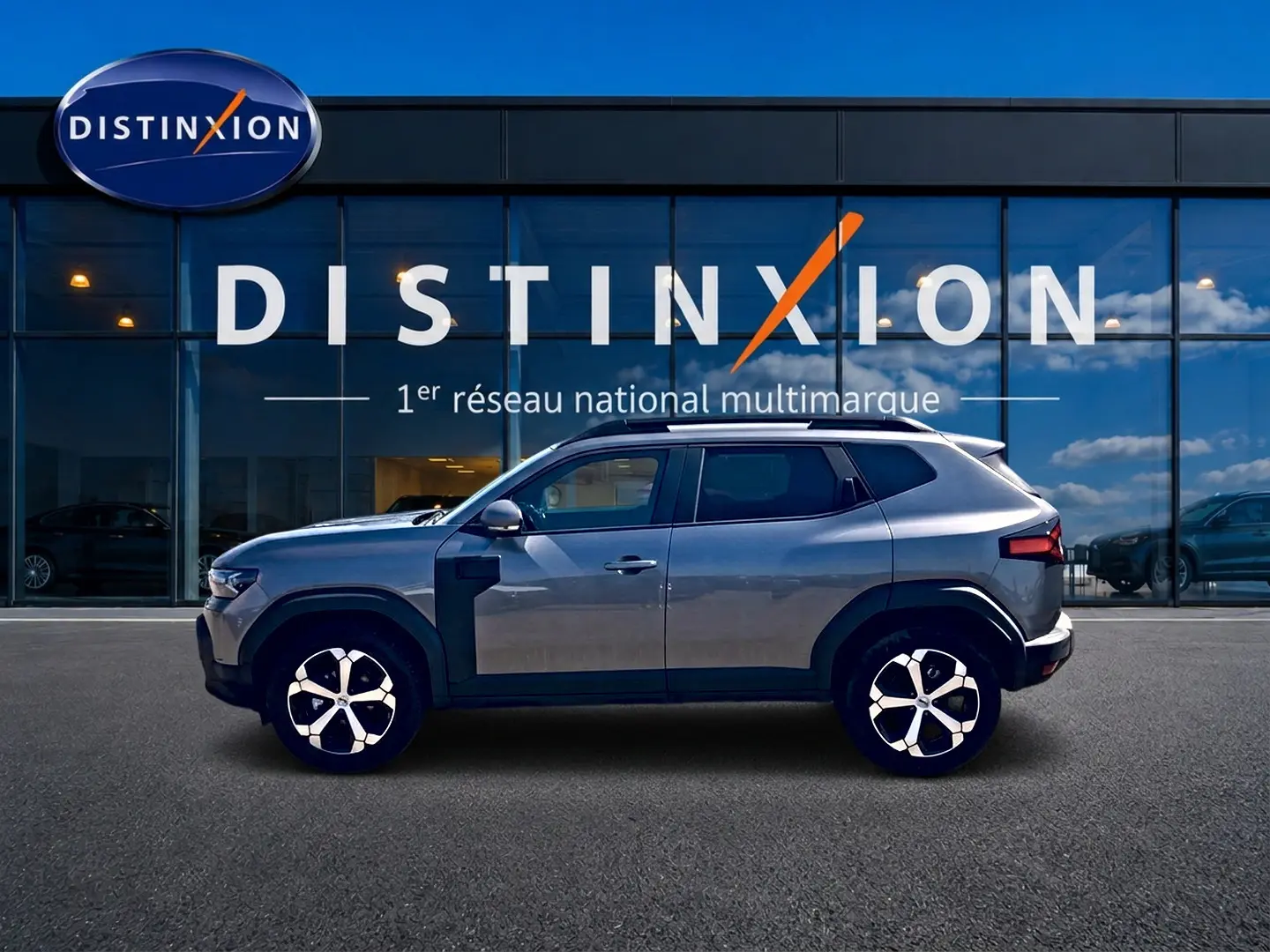 Profil droit du Dacia Duster gris schiste 2026 avec jantes alu et barres de toit, devant une vitrine Distinxion.
