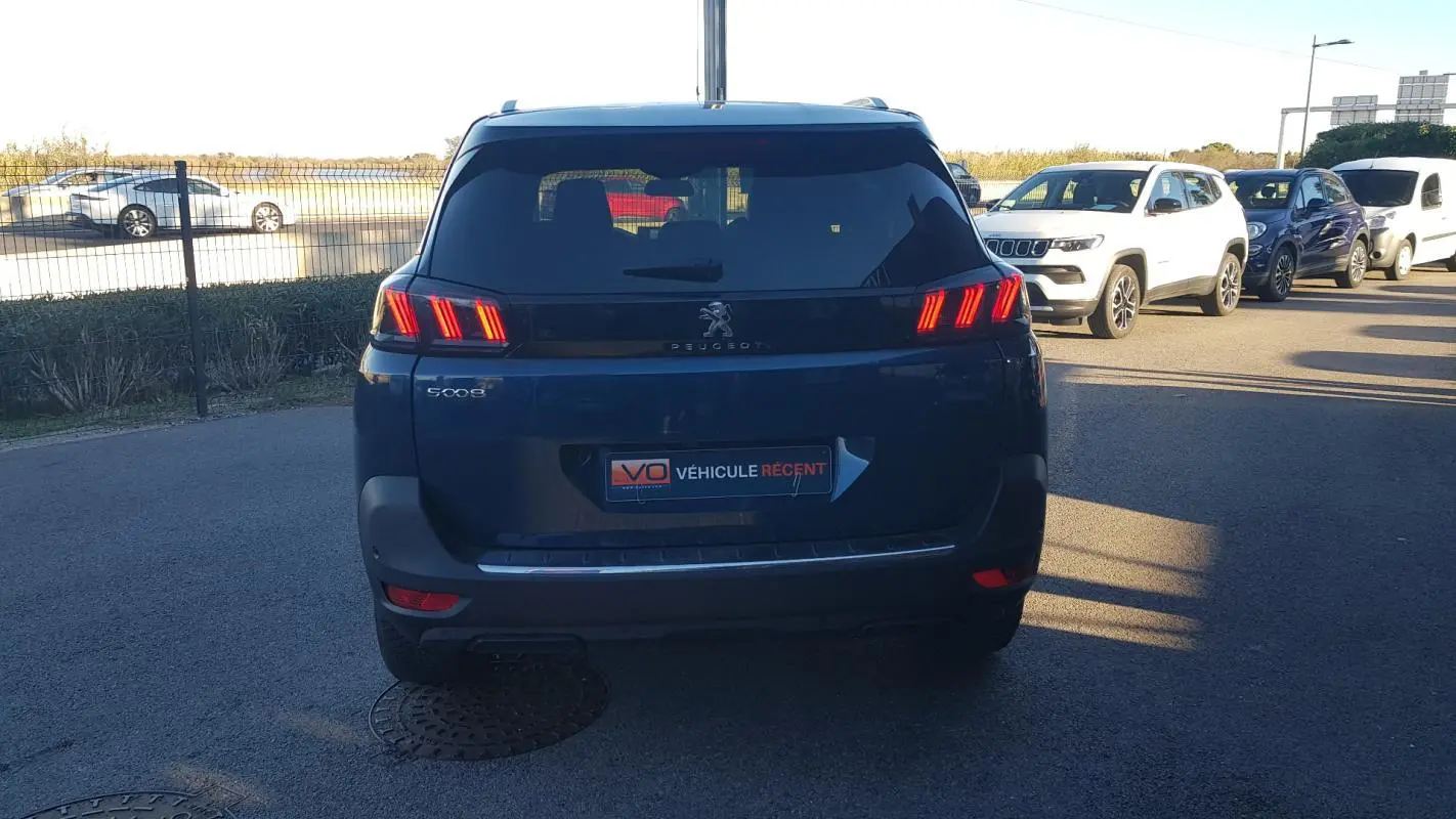Vue arrière d’un Peugeot 5008 bleu 2024 avec feux allumés et barres de toit visibles en extérieur.