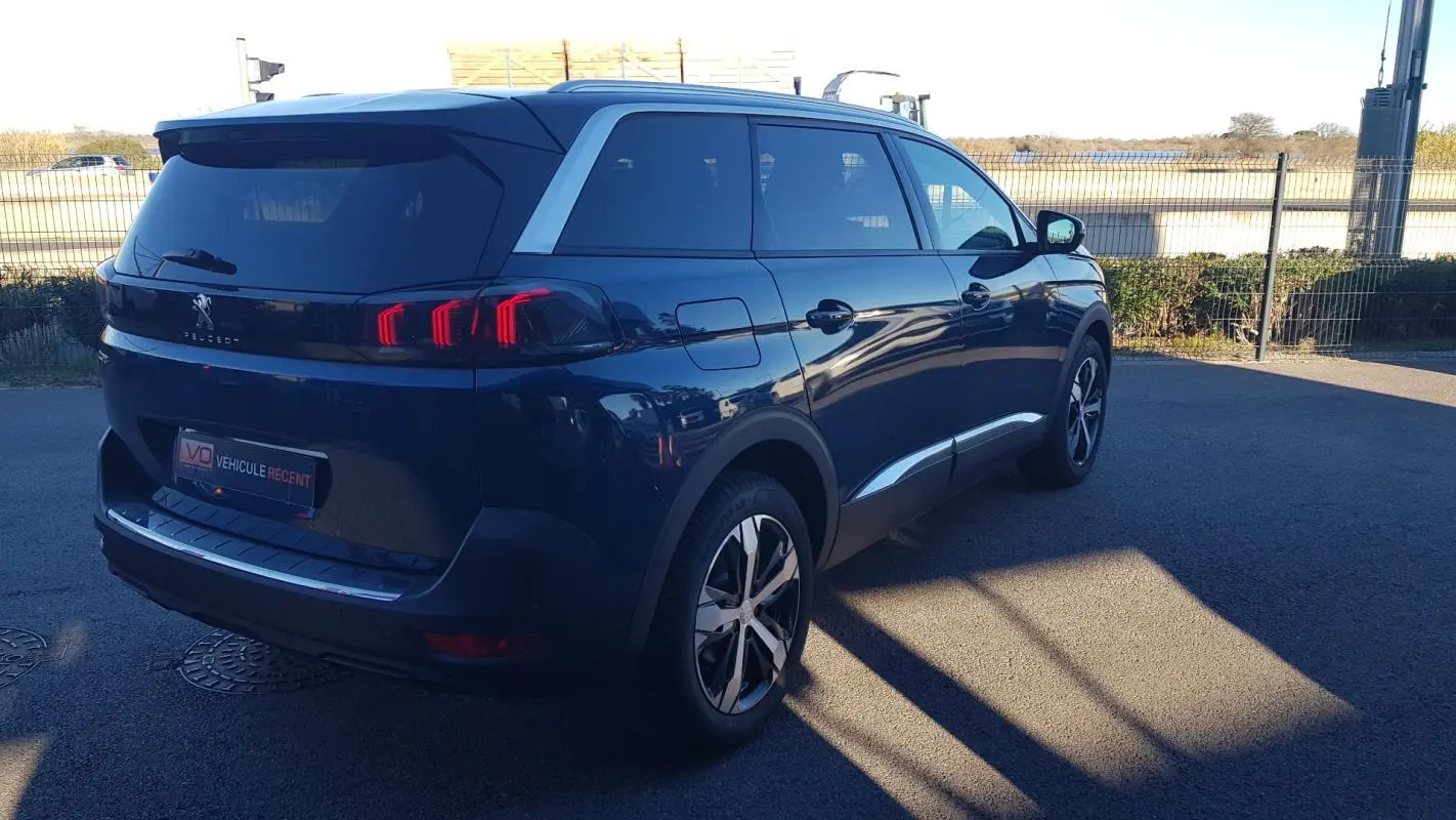 Vue 3/4 arrière droite d’un Peugeot 5008 bleu 2024 avec jantes alliage 18 pouces et barres de toit visibles.