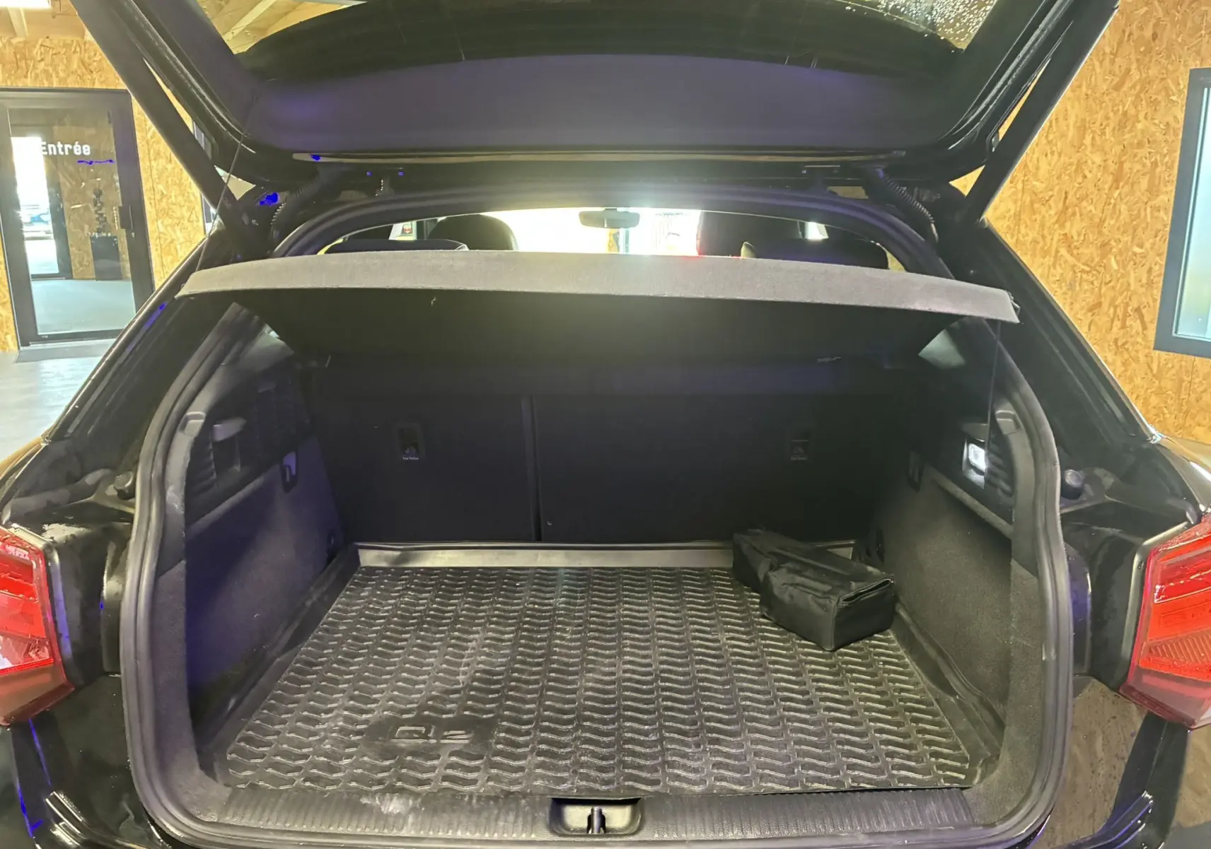 Coffre ouvert d’un Audi Q2 noir vue arrière, avec tapis de coffre et cache bagages partiellement relevé.