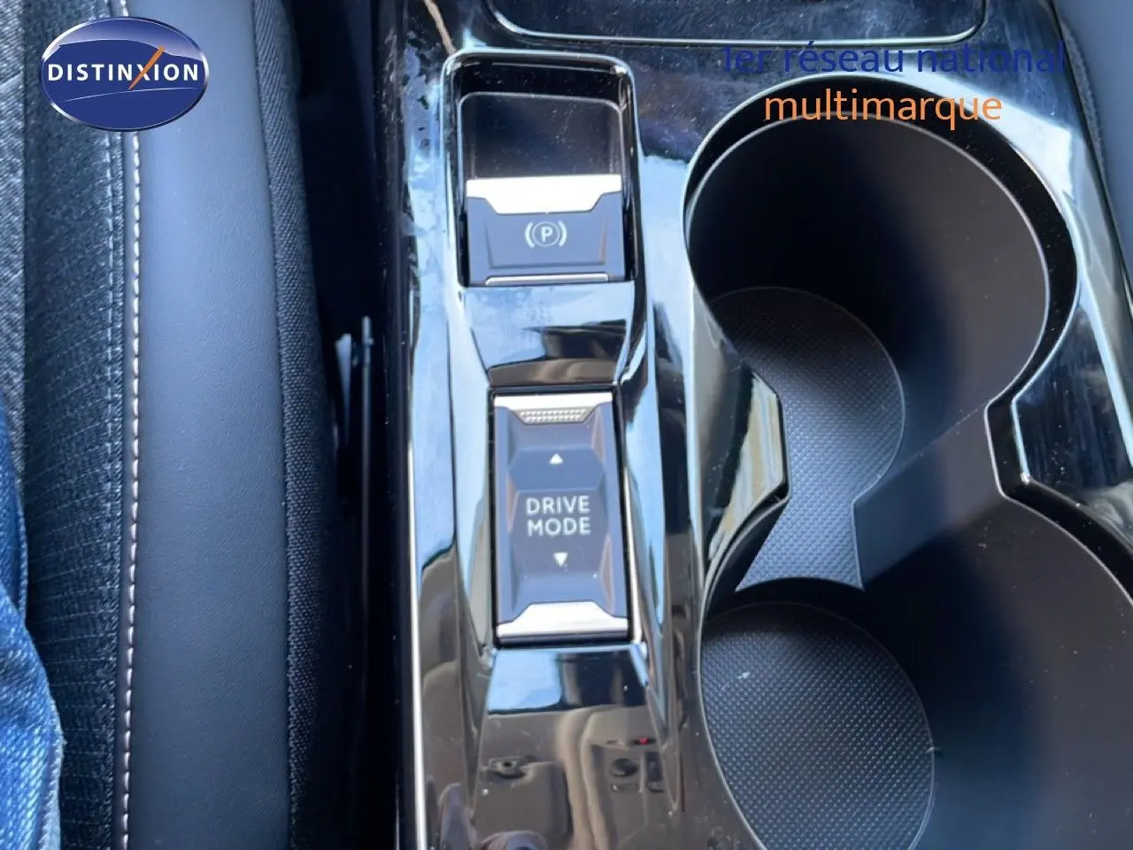 Gros plan sur la console centrale noire brillante avec boutons Drive Mode et frein à main électrique, porte-gobelets visibles.
