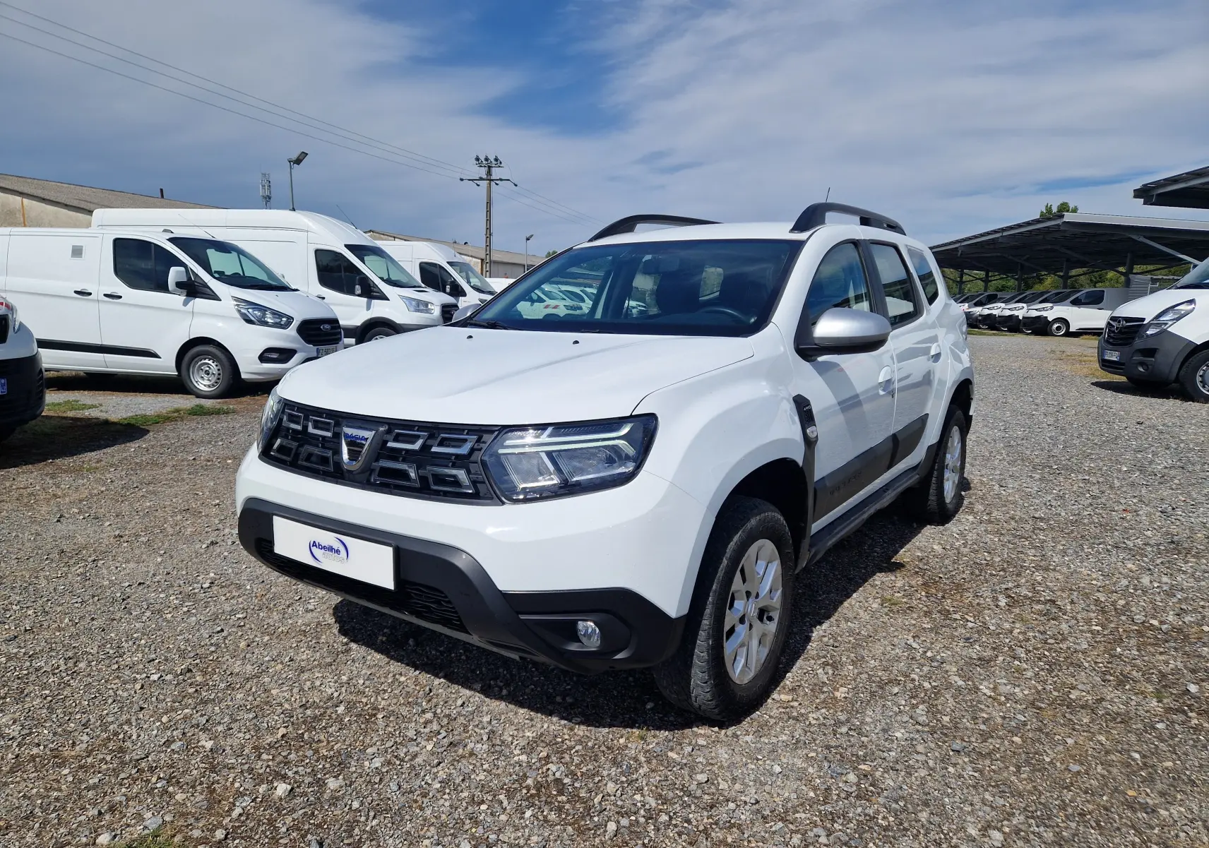 Dacia Duster blanc 4x4 en 3/4 avant droit, avec protections noires sur les portières et calandre distinctive.