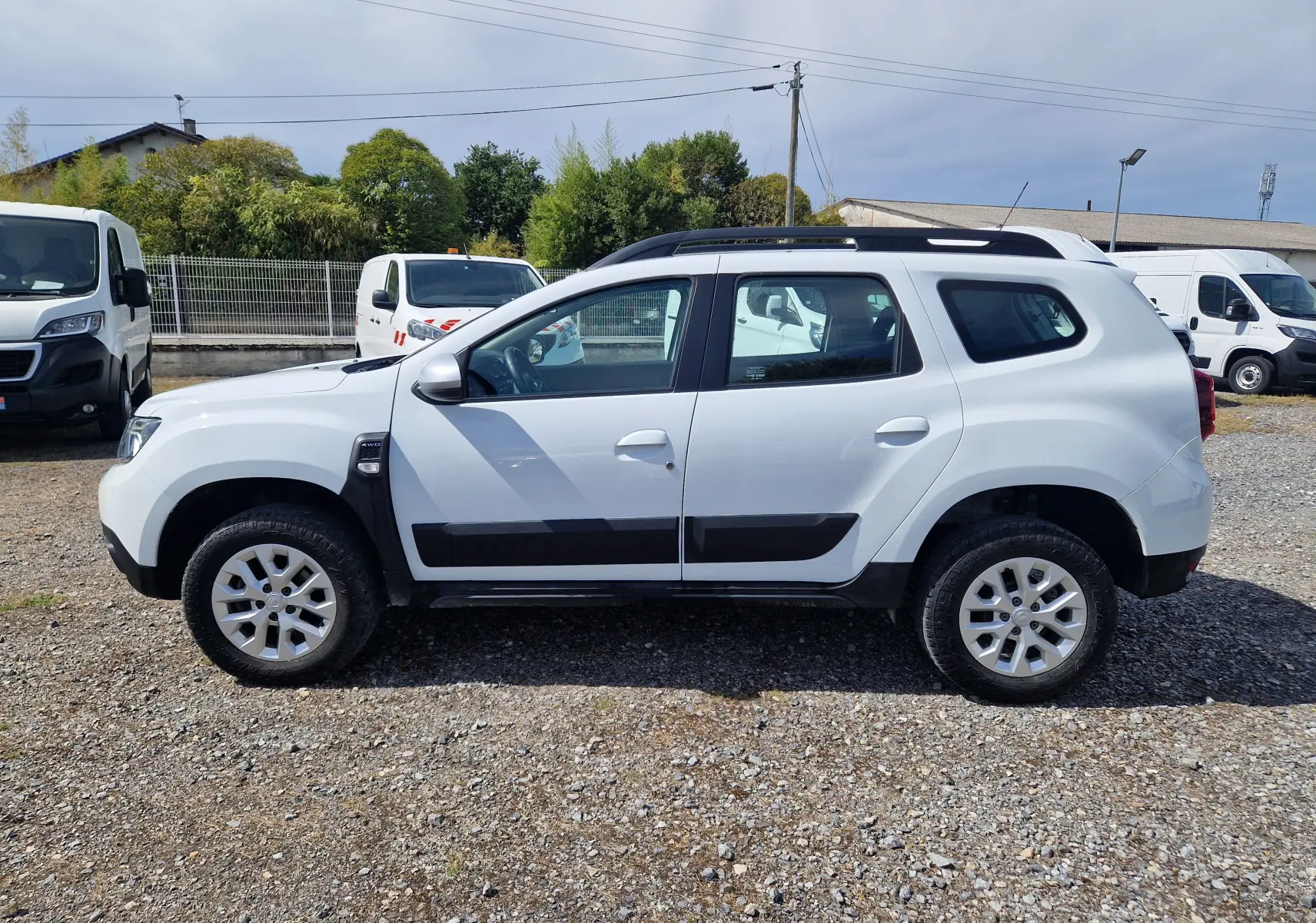 Profil côté gauche d'un Dacia Duster blanc 4x4 2023, avec protections noires sur les portières et jantes alliage 16 pouces.
